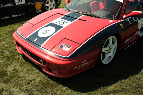 A red sports car with racing decals and the number 18 displayed prominently on its hood. The car features white rims and is parked on a grassy area. Several sponsor logos, including Shell and Girard-Perregaux, are visible on the car's body.