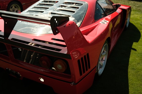 A sports car with a sleek, aerodynamic design is positioned on a grassy field. The car is bright red with a large rear spoiler and visible engine components through a transparent cover. It has angular, aggressive lines and features distinctive rear tail lights.