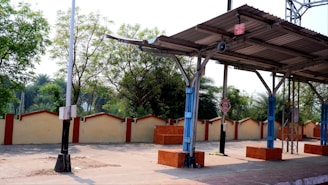 A deserted railway platform with metal roofing, supported by blue and grey pillars. Surrounding the platform are trees and a boundary wall with a cream and red-tone aesthetic. There is a signpost on the platform, and natural light illuminates the scene.