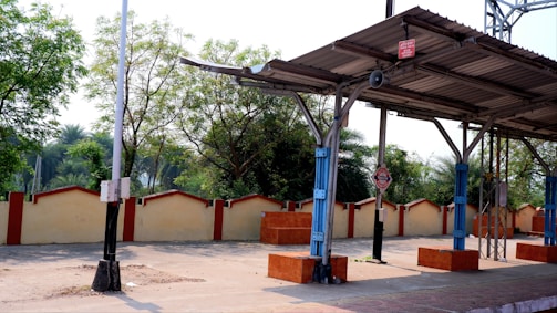 A deserted railway platform with metal roofing, supported by blue and grey pillars. Surrounding the platform are trees and a boundary wall with a cream and red-tone aesthetic. There is a signpost on the platform, and natural light illuminates the scene.