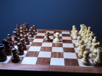 A wooden chess board is set up for a game with dark and light pieces in their starting positions. The chess pieces are made of polished wood, giving a classic and elegant appearance. The board itself has alternating light and dark squares, with a warm, glossy finish.