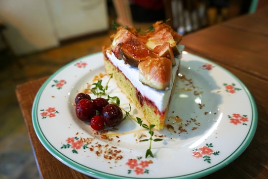 A slice of cake topped with fresh figs and served on a floral patterned plate. The cake has a light, creamy layer with fruit, accompanied by a small cluster of red berries and sprigs of herbs. The setting suggests a cozy, rustic atmosphere.