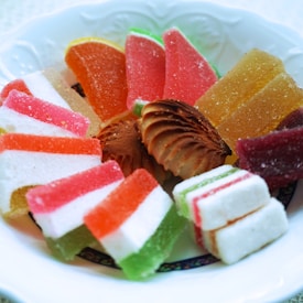 A plate filled with a colorful assortment of sweets, including sugar-coated jelly candies in various shapes and colors and a small cream-filled pastry in the center. The candies are arranged in an aesthetically pleasing manner, with bright hues of pink, orange, red, and green.