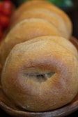 A close-up view of freshly baked buns arranged in a wooden bowl. The buns have a golden-brown crust with a slightly rough texture, indicating they are likely made from whole grains. The background is slightly blurred, emphasizing the focus on the bread.