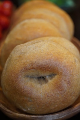 A close-up view of freshly baked buns arranged in a wooden bowl. The buns have a golden-brown crust with a slightly rough texture, indicating they are likely made from whole grains. The background is slightly blurred, emphasizing the focus on the bread.