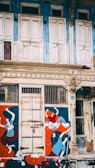 An old, weathered building facade with ornate architectural details. The upper part features closed, white wooden shutters set against a faded blue wall. Below, a door is flanked by vibrant murals of abstract human figures painted in red, blue, and orange colors.