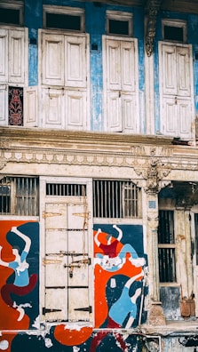 An old, weathered building facade with ornate architectural details. The upper part features closed, white wooden shutters set against a faded blue wall. Below, a door is flanked by vibrant murals of abstract human figures painted in red, blue, and orange colors.