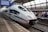 A sleek, modern ICE train is stationed at a platform with a group of passengers waiting nearby. The platform is covered by a large glass roof and a digital sign displays travel information indicating a route to Frankfurt. The train is white with red and blue stripes, and the railway tracks are visible in the foreground.
