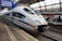A sleek, modern ICE train is stationed at a platform with a group of passengers waiting nearby. The platform is covered by a large glass roof and a digital sign displays travel information indicating a route to Frankfurt. The train is white with red and blue stripes, and the railway tracks are visible in the foreground.