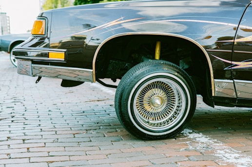 A classic car with a raised wheel, emphasizing its detailed wire-spoke rims and a shiny exterior. The car is parked on cobblestone pavement, suggesting an outdoor or urban setting.