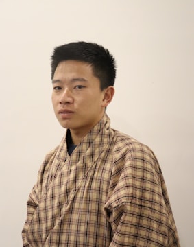 A person is wearing a traditional checkered garment with a neutral expression. The background is plain and light-colored, emphasizing the attire and the person's features.