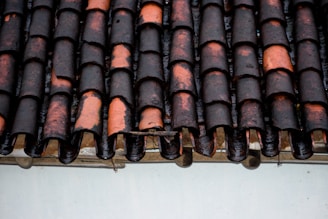 A close-up view of a weathered, traditional terracotta roof with overlapping curved tiles. The tiles show signs of wear and some areas are darker, possibly indicating moisture or the development of moss or dirt.