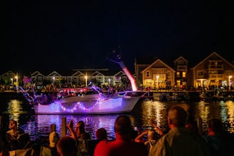 A boat adorned with bright, colorful lights sails on a calm body of water at night. In the background, a crowd of people is gathered along the shoreline, watching the illuminated spectacle. A row of houses is visible, with warm lights glowing from the windows. The American flag prominently decorates the boat.