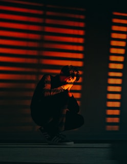 A person is crouched on the floor, with hands covering their face, under dramatic lighting that casts orange-red striped shadows across the scene. The environment is dark with prominent contrasts created by the lines of light and shadow.