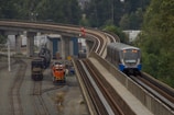 A modern passenger train travels along an elevated railway track beside a rail yard. The rail yard contains two stationary freight locomotives and several tracks. The area is surrounded by green trees and industrial structures.