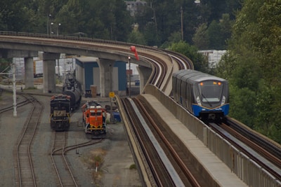 A modern passenger train travels along an elevated railway track beside a rail yard. The rail yard contains two stationary freight locomotives and several tracks. The area is surrounded by green trees and industrial structures.