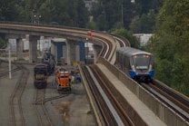 A modern passenger train travels along an elevated railway track beside a rail yard. The rail yard contains two stationary freight locomotives and several tracks. The area is surrounded by green trees and industrial structures.