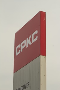 A tall sign with the letters 'CPKC' in bold white font on a red background. Below it, there is a partial view of a grey section with the text 'Vancouver'. The sign has a clean, modern appearance and stands against a cloudy sky.