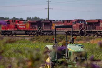 A green tractor is positioned in the foreground with several red locomotives in the background, belonging to Canadian Pacific Railway. The scene takes place in a rural area with green fields and distant trees under a blue sky. Purple flowers are also visible, blurred in the foreground.