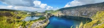 A panoramic view of a large, peaceful quarry lake surrounded by rocky cliffs and lush green vegetation under a bright blue sky filled with fluffy white clouds. In the distance, there is a building nestled among trees, and the landscape stretches into the horizon.