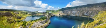 A panoramic view of a large, peaceful quarry lake surrounded by rocky cliffs and lush green vegetation under a bright blue sky filled with fluffy white clouds. In the distance, there is a building nestled among trees, and the landscape stretches into the horizon.