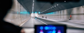 A tunnel interior with multiple vehicles traveling through it. The view is from inside another vehicle, focusing on the road ahead. The tunnel is well-lit with overhead lights, and the walls are smooth and slightly reflective. There are several lanes, with cars and motorcycles visible.