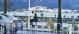 A couple stands closely together near a fence by the waterfront, with one person appearing to check something on a mobile device. In the background, there are people walking along a beach with a well-lit building in the distance. A car is parked to the left, partially obscured by the fence. The setting appears to be in the evening or late afternoon with a relaxed ambiance.