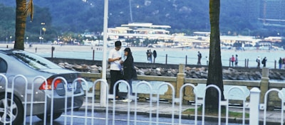 A couple stands closely together near a fence by the waterfront, with one person appearing to check something on a mobile device. In the background, there are people walking along a beach with a well-lit building in the distance. A car is parked to the left, partially obscured by the fence. The setting appears to be in the evening or late afternoon with a relaxed ambiance.