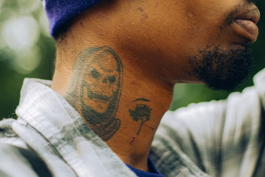 A close-up view of a person's neck with a tattoo depicting a grim reaper. The tattoo features a skull with a hood, inked in dark tones. The person is wearing a plaid shirt and a purple beanie, and their facial hair is visible.