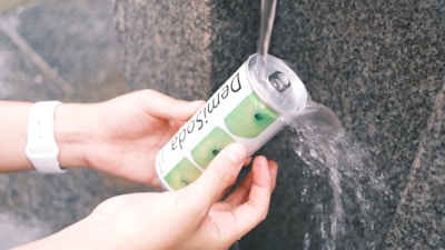 Hands holding a beverage can with water being poured over it from a stream. The can has images of green apples and the word 'Demisoda.' The person is wearing a white wristwatch.
