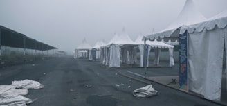 An array of white tents is lined up along an empty road in a misty and overcast setting. The tents appear to be part of an outdoor event or expo, evidenced by banners with the word 'expo' and the year 2023. The area is deserted, with some tents partially set up and others having fallen fabric on the ground. The overall scene is muted and devoid of people.
