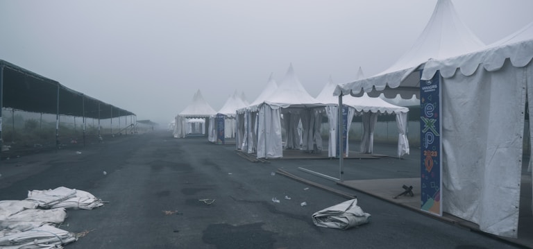 An array of white tents is lined up along an empty road in a misty and overcast setting. The tents appear to be part of an outdoor event or expo, evidenced by banners with the word 'expo' and the year 2023. The area is deserted, with some tents partially set up and others having fallen fabric on the ground. The overall scene is muted and devoid of people.