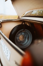 The image depicts the trunk of a vintage car with the lid open, revealing a spare tire inside. The interior of the trunk is beige, and the spare tire is mounted on a metal rim with a rust-free appearance. The scene includes part of a car's license plate visible on the side.