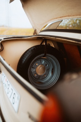 The image depicts the trunk of a vintage car with the lid open, revealing a spare tire inside. The interior of the trunk is beige, and the spare tire is mounted on a metal rim with a rust-free appearance. The scene includes part of a car's license plate visible on the side.