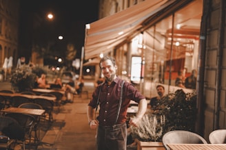 A man with a friendly expression stands confidently outside a warmly lit street cafe at night. Several empty tables and chairs are arranged around him, while a few patrons sit at tables in the background. The setting has a cozy and inviting atmosphere with a mix of outdoor and indoor seating visible through large glass windows.