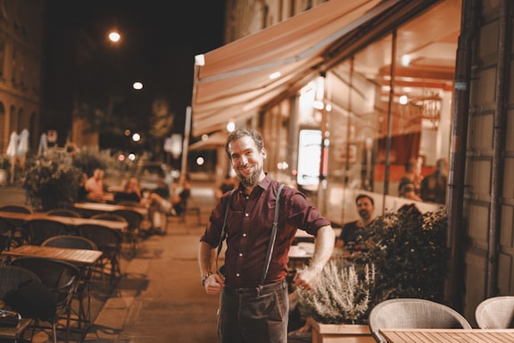 A man with a friendly expression stands confidently outside a warmly lit street cafe at night. Several empty tables and chairs are arranged around him, while a few patrons sit at tables in the background. The setting has a cozy and inviting atmosphere with a mix of outdoor and indoor seating visible through large glass windows.