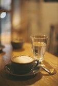 A warm, cozy atmosphere with a cup of frothy cappuccino in a ceramic mug and saucer on a wooden table, accompanied by a glass of water. The blurred background suggests a relaxed coffee shop setting.