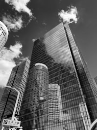 A group of tall skyscrapers with glass facades reflects the sky and clouds above. The architecture is modern and imposing, with clean lines and a sleek design. There is a street sign that indicates directions to Osaka City.