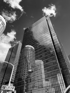 A group of tall skyscrapers with glass facades reflects the sky and clouds above. The architecture is modern and imposing, with clean lines and a sleek design. There is a street sign that indicates directions to Osaka City.