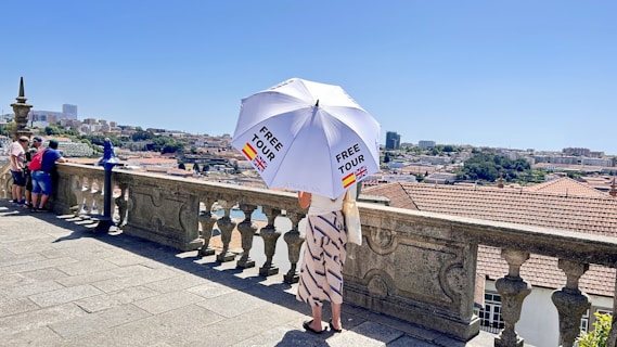 A person stands on a stone terrace overlooking a cityscape, holding a large white umbrella with 'FREE TOUR' printed on it and flags of Spain and the UK. The terrace has a decorative stone balustrade, and several other people are visible gazing at the view. The background shows a city with buildings and greenery under a clear blue sky.