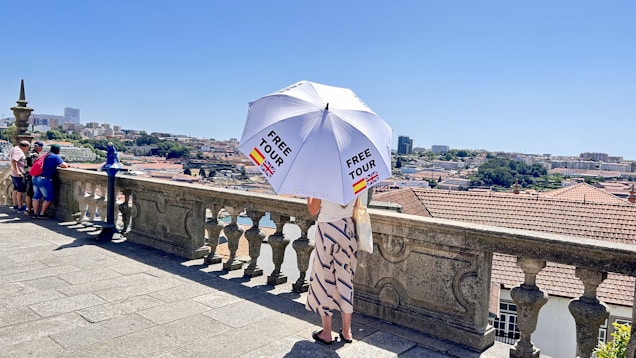 A person stands on a stone terrace overlooking a cityscape, holding a large white umbrella with 'FREE TOUR' printed on it and flags of Spain and the UK. The terrace has a decorative stone balustrade, and several other people are visible gazing at the view. The background shows a city with buildings and greenery under a clear blue sky.