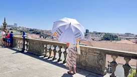 A person stands on a stone terrace overlooking a cityscape, holding a large white umbrella with 'FREE TOUR' printed on it and flags of Spain and the UK. The terrace has a decorative stone balustrade, and several other people are visible gazing at the view. The background shows a city with buildings and greenery under a clear blue sky.