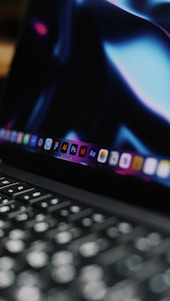 A close-up view of a laptop keyboard with the screen displaying various software icons. The screen has a dark background with bluish and purplish hues.