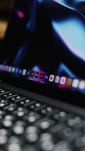 A close-up view of a laptop keyboard with the screen displaying various software icons. The screen has a dark background with bluish and purplish hues.