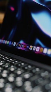 A close-up view of a laptop keyboard with the screen displaying various software icons. The screen has a dark background with bluish and purplish hues.