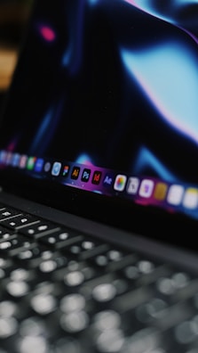 A close-up view of a laptop keyboard with the screen displaying various software icons. The screen has a dark background with bluish and purplish hues.