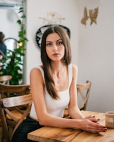 A woman with long brown hair and a serious expression sits at a wooden table in a well-lit room. She wears a white sleeveless top and has her hands resting gently on the table. The background features wooden chairs, potted plants, and decorative elements on the walls.