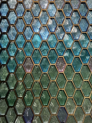 A pattern of hexagonal tiles in shades of blue and green, each with a textured, diamond-like interior design. The tiles are arranged in a regular, grid-like pattern, creating a visually striking and geometric display.