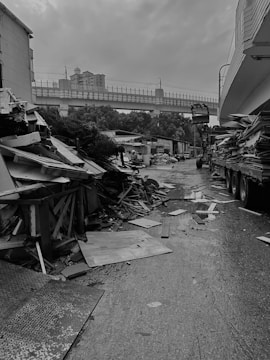 A street lined with debris and construction materials, including large piles of wooden planks and panels. The road appears wet, and the atmosphere is overcast. In the background, there is an elevated railway with a train track and a couple of tall buildings.