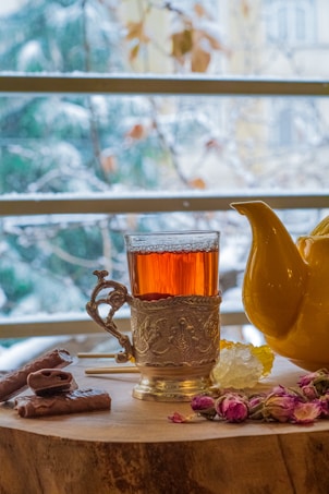 A glass of tea with an ornate metal holder sits on a wooden surface. Beside it is a yellow teapot. The scene is embellished with cinnamon sticks, a piece of crystal sugar, and some dried rosebuds. Outside the window in the background, snow-covered trees and branches are visible.
